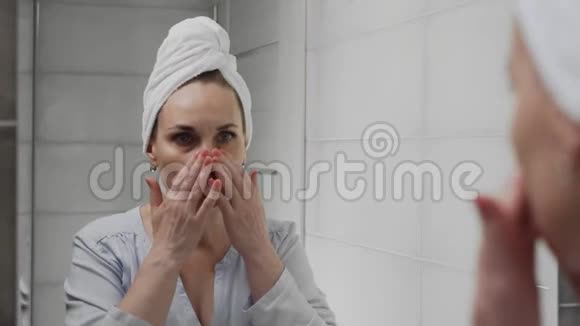 沮丧的成年女人头上戴着毛巾在浴室镜子里看着她的脸视频的预览图