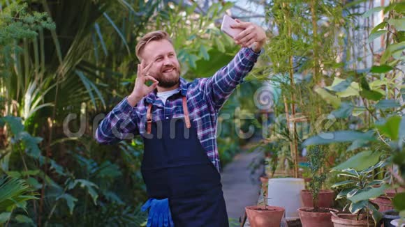 快乐和微笑的大园丁男人用智能手机在大温室里自拍视频的预览图