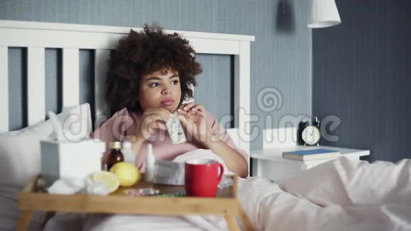 生病的非裔美国女孩在家里躺在床上用水吃药视频的预览图