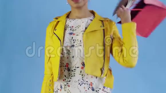 美国黑人妇女带着购物袋和手机在蓝底工作室的肖像黄色夹克视频的预览图