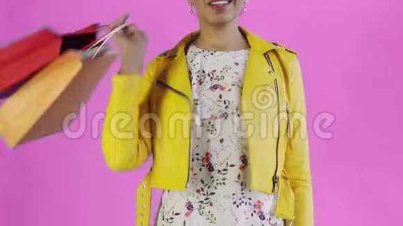 美国黑人妇女在工作室的粉红色背景上带购物袋的肖像幸福的女人手里拿着视频的预览图