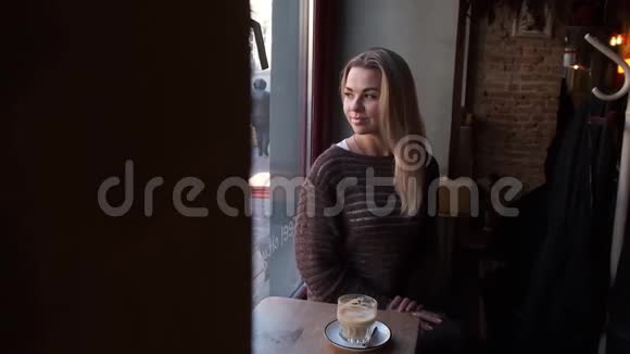 年轻漂亮的女人留着长长的金发坐在咖啡馆的窗户附近喝着咖啡视频的预览图