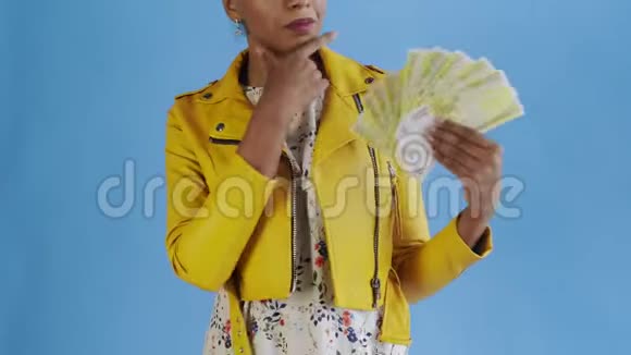 深思熟虑的成功的非裔美国妇女与金钱迷蓝背景在工作室黄色夹克视频的预览图