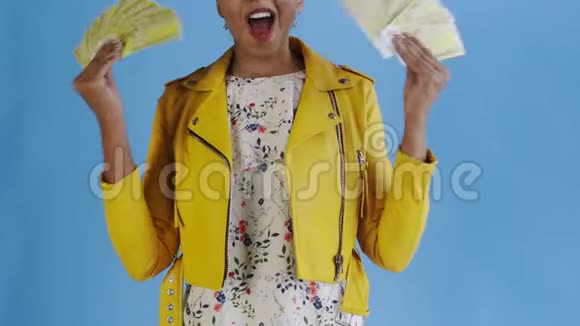 高兴兴奋的非裔美国妇女在蓝色背景的黄色夹克上炫耀金钱说上帝保佑视频的预览图