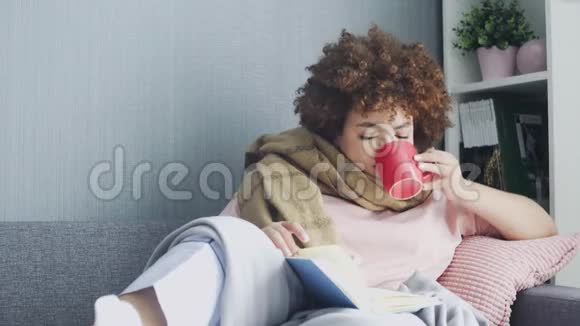 一个女人家里感冒了躺在沙发上拿着一本书和一杯热茶上面盖着一条毯子视频的预览图