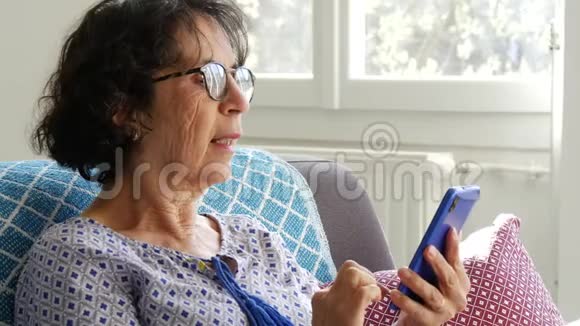 黑发开朗的高级女性坐在沙发上用智能手机视频的预览图