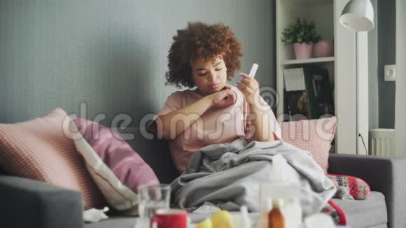 生病的非洲裔美国流感女孩躺在家里的沙发上病了的年轻黑人妇女用感冒检查体温视频的预览图