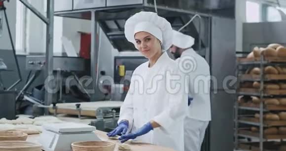可爱的成熟女人带着笑容的大面包师穿着白色制服准备面团烘焙面包而其他工人视频的预览图