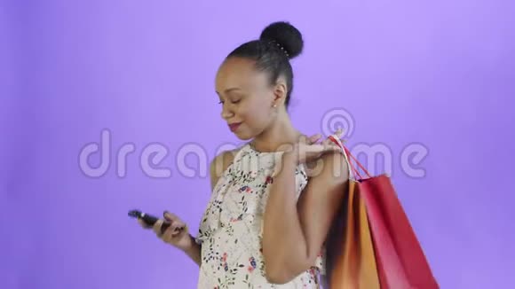 美国黑人妇女带着购物袋的肖像在工作室的紫色背景上通过智能手机交谈白色视频的预览图