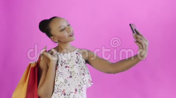 一位拿着购物袋的非裔美国妇女正在工作室里用粉色背景自拍带花的白色连衣裙视频的预览图