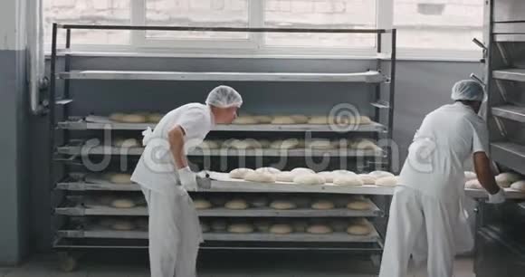 在两个面包店工人的烤箱机器中装载生面包的过程在摄像机前走来走去视频的预览图