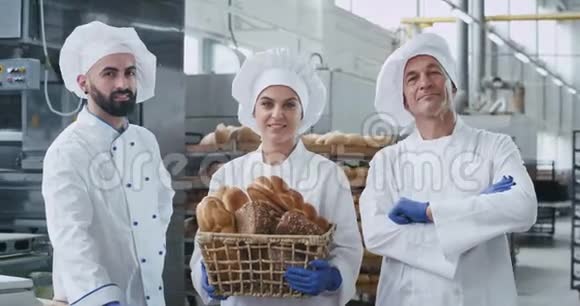 在镜头前微笑着三个穿着制服的面包师在一家大面包店拿着一个装有有机食品的老式篮子视频的预览图