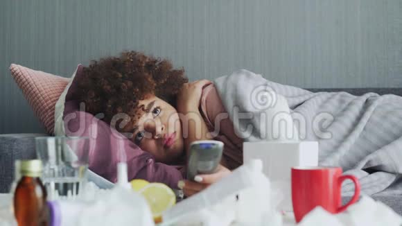 生病的非裔美国女孩在家里患流感生病的年轻黑人妇女发烧看电视节目和使用遥控器视频的预览图