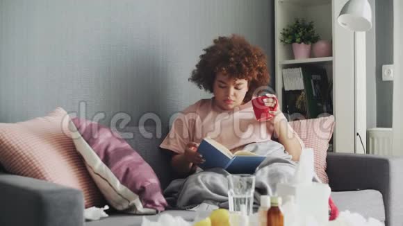 一个女人家里感冒了躺在沙发上拿着一本书和一杯热茶上面盖着一条毯子视频的预览图