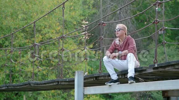 迷人英俊的金发男子坐在河上的吊桥上风景如画视频的预览图