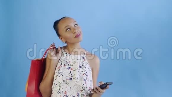 年轻美丽的非洲人使用电话和拿着购物袋在蓝色背景在工作室带花的白色裙子视频的预览图