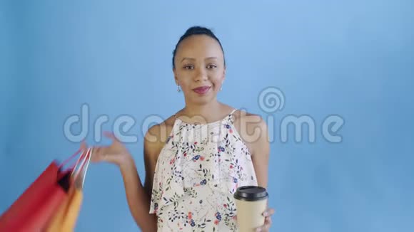 美国黑人妇女带着购物袋的肖像正在工作室的蓝色背景上喝咖啡J白色连衣裙视频的预览图