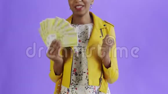 满意的兴奋的兴奋的非裔美国女人在紫色背景的黄色夹克上显示金钱和说YEAH视频的预览图