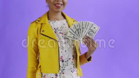美国黑人妇女在工作室的紫色背景上带着钱迷的肖像黄色夹克视频的预览图
