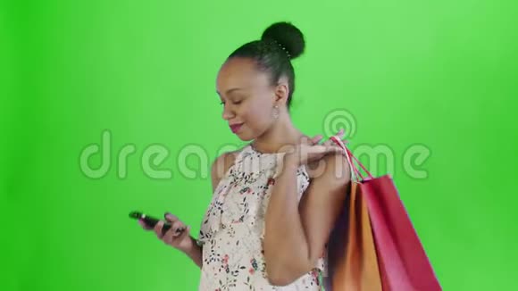 美国黑人妇女带着购物袋的肖像是在工作室的绿色背景上通过智能手机谈论的白色视频的预览图