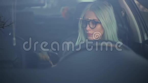 年轻活泼的金发司机女孩手里拿着智能手机戴着太阳镜和牛仔裤夹克在车窗上的肖像视频的预览图