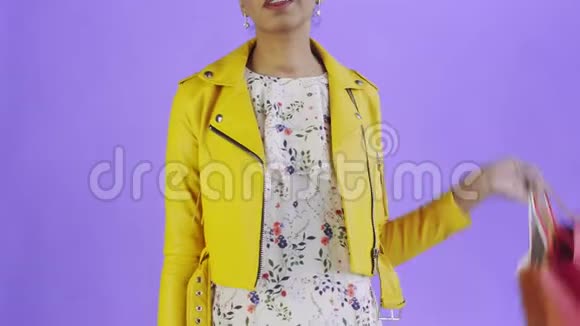 非洲裔美国妇女的肖像与购物袋和黄金信用卡在紫色背景在工作室黄色夹克视频的预览图