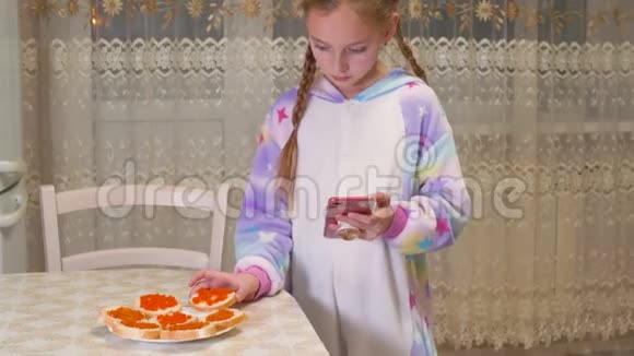 可爱的小女孩使用智能手机在家吃红鱼子酱视频的预览图