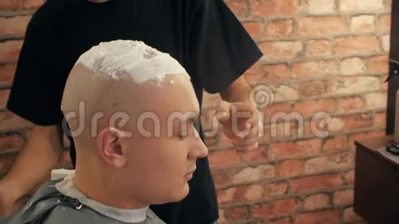 理发店里一个年轻的秃头正拿着剃须刀理发师用剃刀和肥皂剃光头视频的预览图