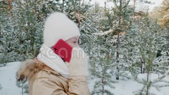 戴着白色帽子和手套的漂亮女人在雪地森林里散步冬天打手机微笑的女人说话视频的预览图