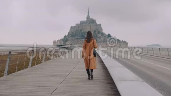 镜头跟随美丽快乐的女人走在史诗山圣米歇尔要塞岛上灰色阴天慢动作视频的预览图