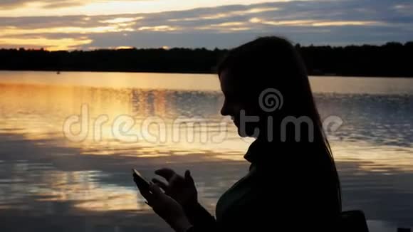 傍晚日落时湖上女游客剪影使用智能手机上网视频的预览图
