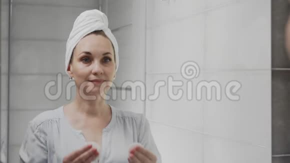 成年女性头上有一条毛巾涂抹乳液照镜子享受自然的美丽和健康视频的预览图