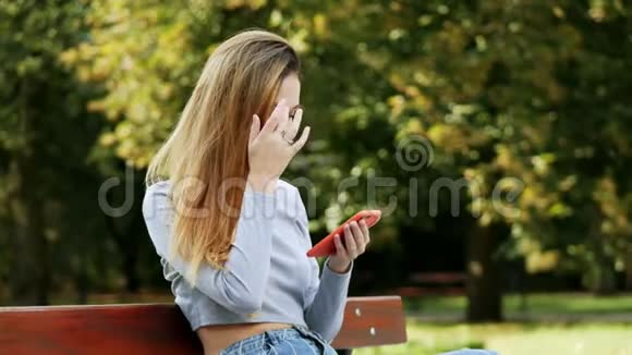 可爱的红发年轻女子在公园的长凳上用智能手机年轻女子一边上网一边微笑视频的预览图