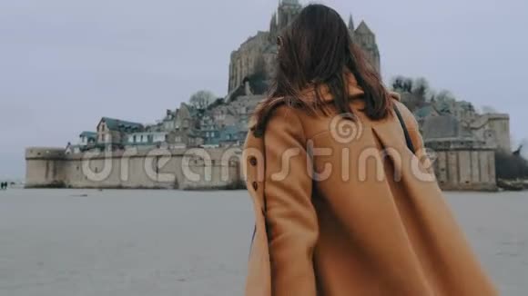 后景特写时尚博主身穿优雅外套步行至圣米歇尔山城堡地标性慢行视频的预览图