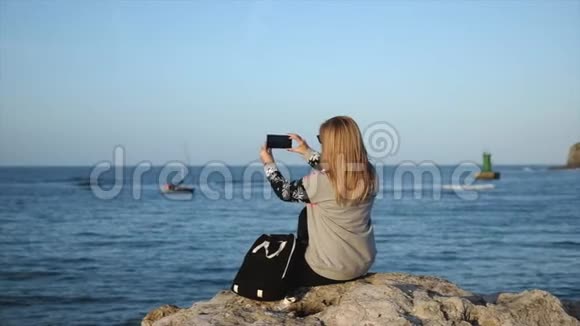 坐在海边的石墙上的女人拍下了一张在阳光明媚的日子里航行的帆船的照片视频的预览图