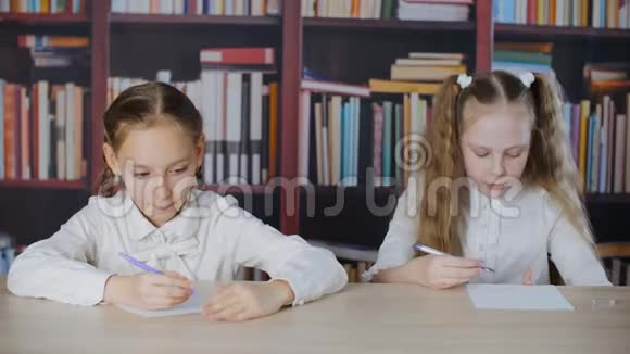 在书架背景下女学生在学校考试中从作弊单上写作两个学生女孩解决作弊问题视频的预览图