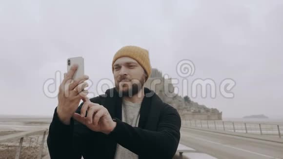快乐年轻兴奋的白种人博客男子拍摄智能手机自拍视频史诗山圣米歇尔城堡慢动作视频的预览图