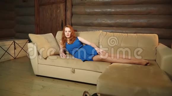 那个漂亮的顽皮的女孩穿着蓝色连衣裙躺在皮革沙发上在木屋里调情和微笑视频的预览图