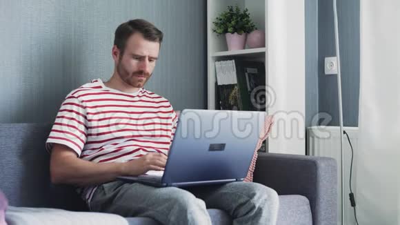 沮丧和疲惫的年轻人在笔记本电脑上工作把它放在膝上坐在家里的沙发上视频的预览图