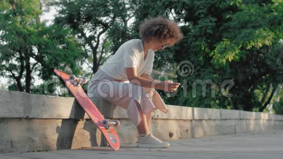 一位坐在公园里玩滑板和打电话的时髦年轻女子的肖像视频的预览图