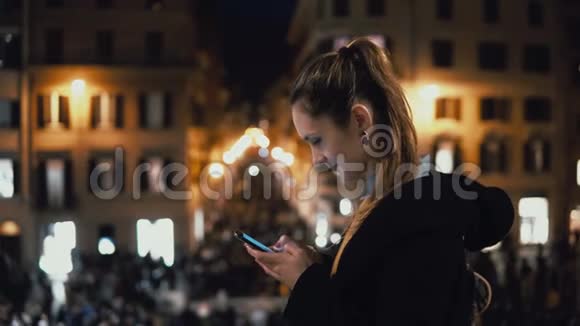 年轻迷人的女人晚上站在市中心使用智能手机背景上的人群和灯光视频的预览图