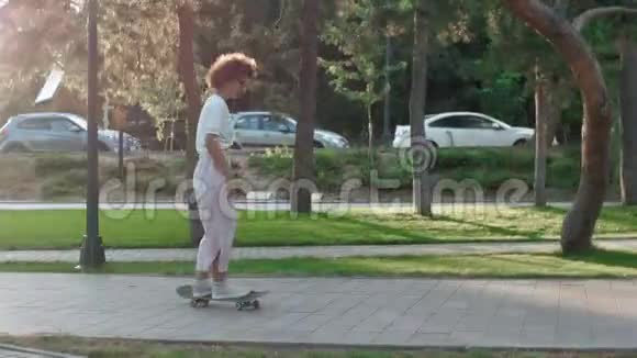 美丽的年轻女孩在公园日落时卷发骑在滑板上视频的预览图