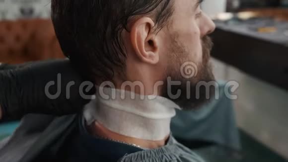 理发师在男沙龙给留胡子的男人理发前穿上披风理发店发型师与客户合作视频的预览图