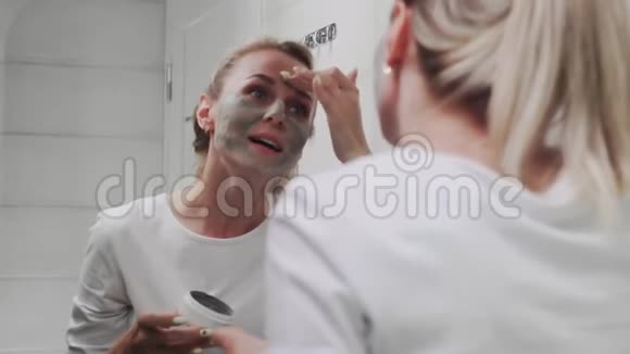 一位妇女在镜子前给自己的脸上涂上粘土面膜视频的预览图