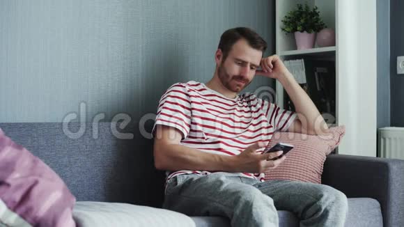 英俊的男人在家里坐在沙发上同时使用智能手机做各种触摸和滑动手势男子视频的预览图