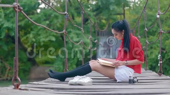 美丽的黑发可爱的女孩坐在河上的吊桥上看书视频的预览图