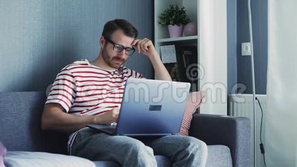 疲劳的年轻人戴着眼镜坐在家里的沙发上在笔记本电脑上工作视频的预览图