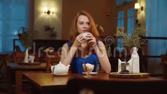 这位美丽的性感女孩红头发穿着蓝色裙子坐在咖啡馆里喝着白色杯子里的茶微笑着视频的预览图