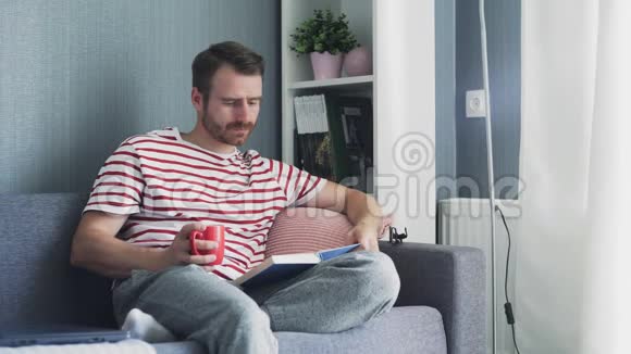 年轻人坐在客厅的沙发上用茶或咖啡杯看书视频的预览图
