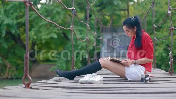 美丽的黑发可爱的女孩坐在河上的吊桥上看书视频的预览图